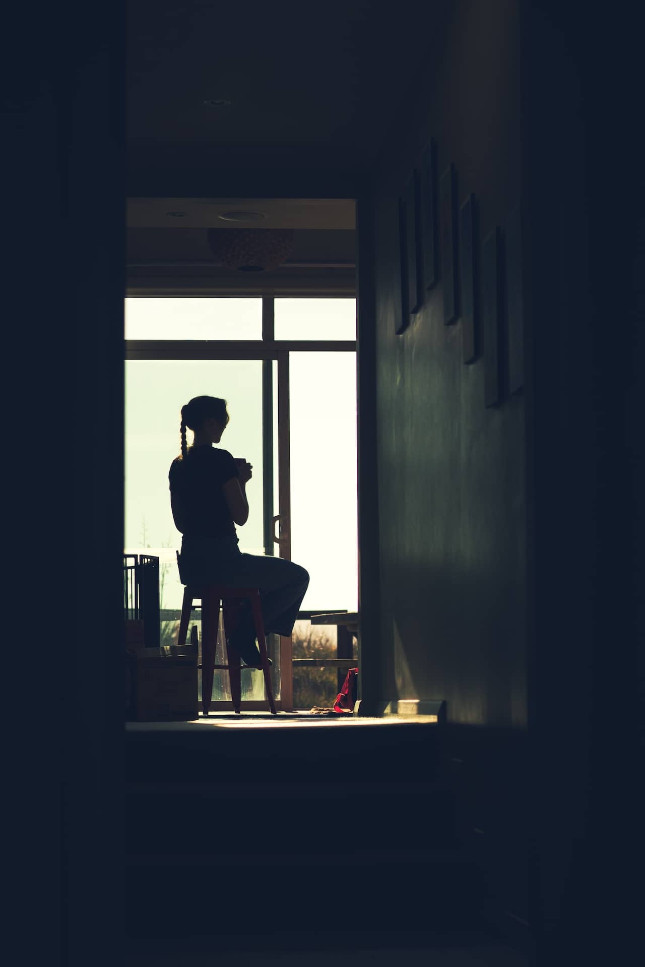 Silhouette photo of a tall woman sipping coffee on a chair, taken through a hallway with the subject framed by the window.